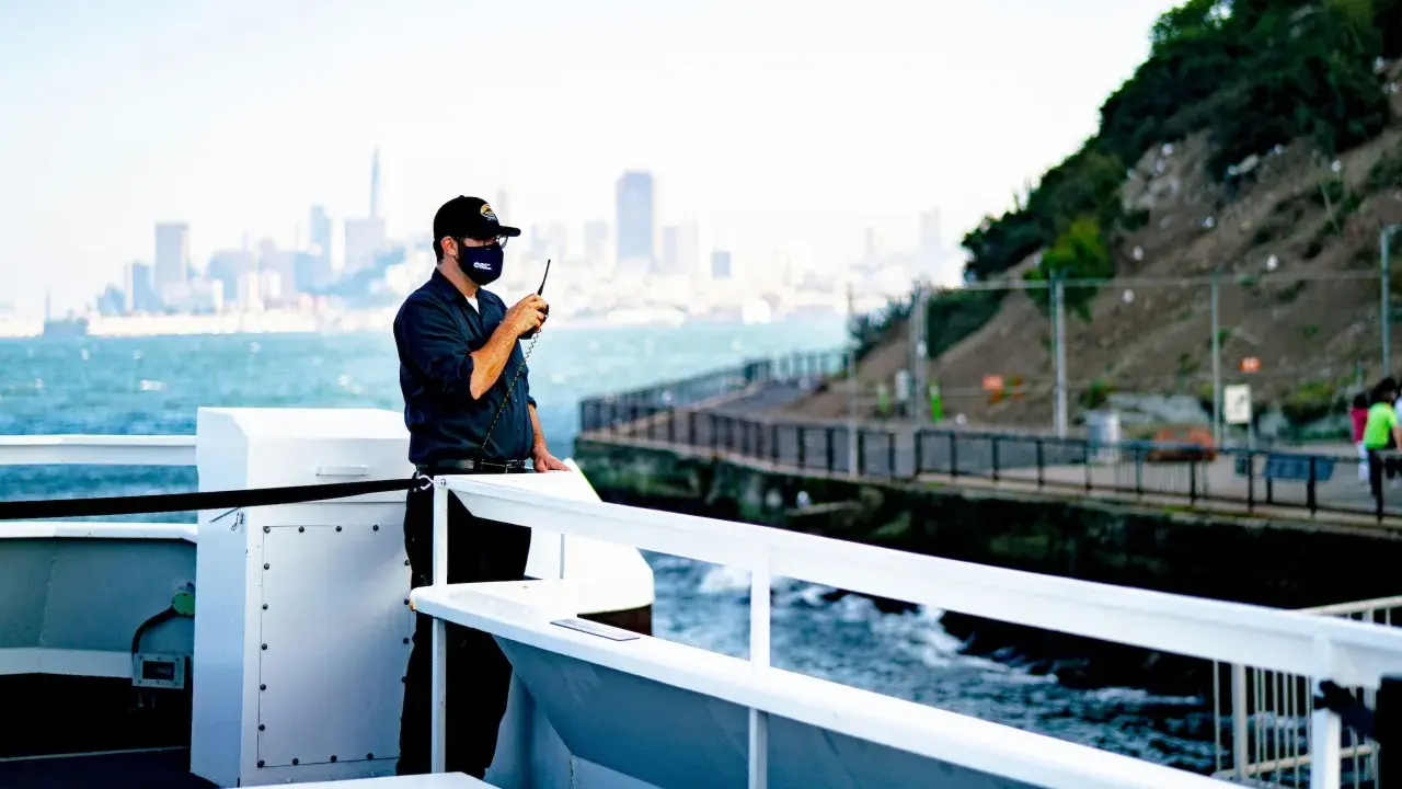 Un homme sur un bateau utilise une radio. Il est en communication, peut-être sur une fréquence vhf libre, avec la ville en arrière-plan.