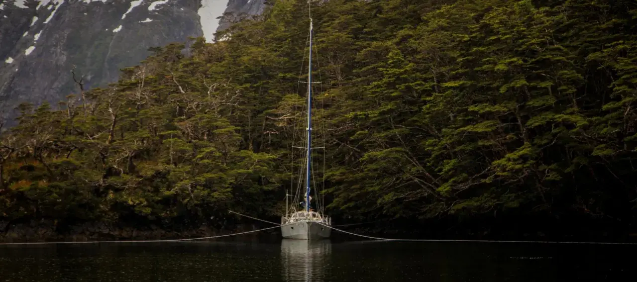 Voilier ancré dans un fjord, entouré d'une forêt dense. La chaîne du cablot, avec son épissure à 3 torons, assure la stabilité.