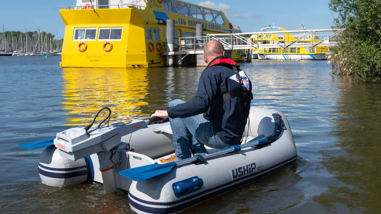 Un homme pilote un bateau gonflable propulsé par la puissance d'un moteur électrique, naviguant près d'un grand bateau jaune.