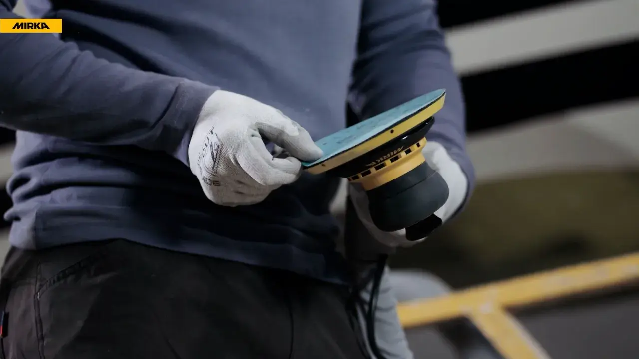 Gants blancs ajustant un disque sur une ponceuse Mirka, prêt à polir la coque d'un bateau.