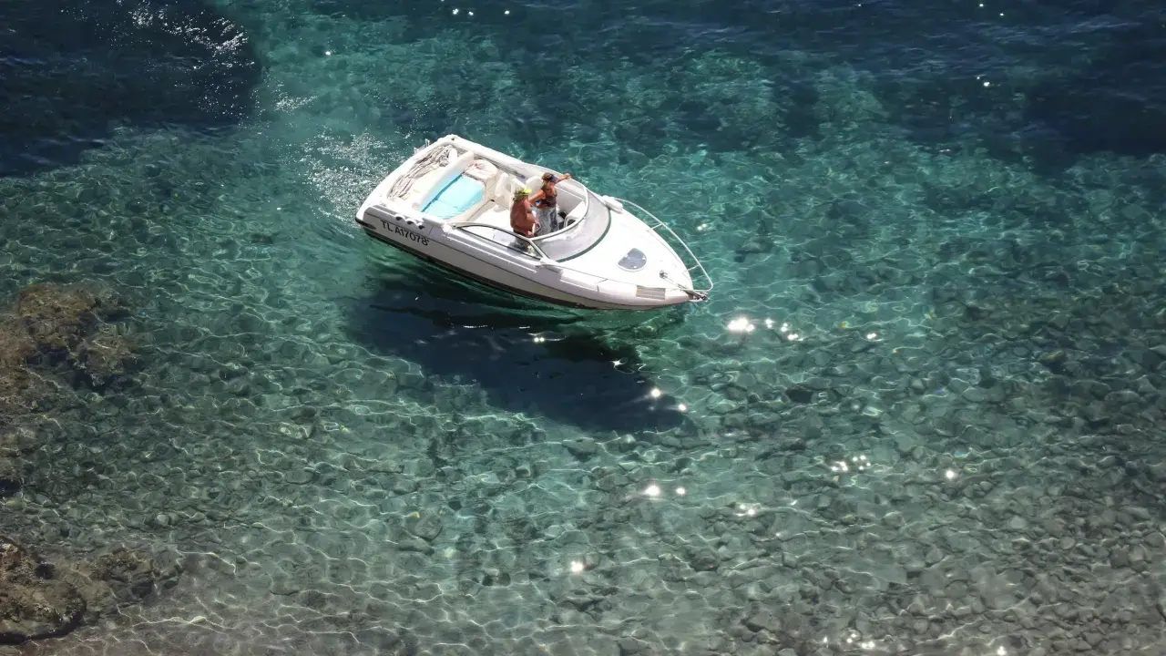 Deux personnes dans un bateau blanc, au moteur en panne, dérivent sur une eau turquoise et claire, révélant les fonds rocheux.