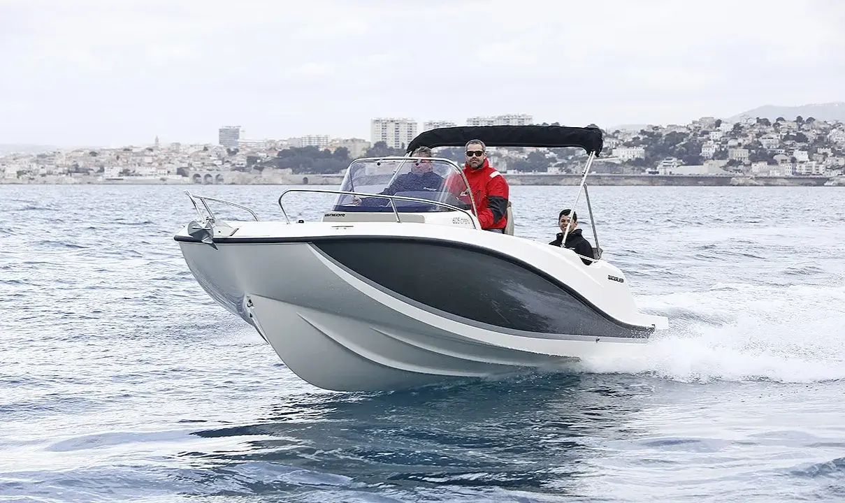 Bateau blanc et gris naviguant sur l'eau, avec trois personnes à bord. Le tableau de bord indique la puissance du moteur du bateau. Ville en arrière-plan.