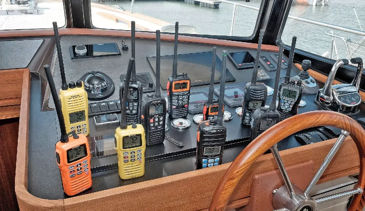 Plusieurs talkies-walkies sur un tableau de bord de bateau. Ils sont de différentes couleurs (orange, jaune, noir) et affichent des canaux. Une discussion sur la différence entre UHF et VHF pourrait être animée avec ces appareils.