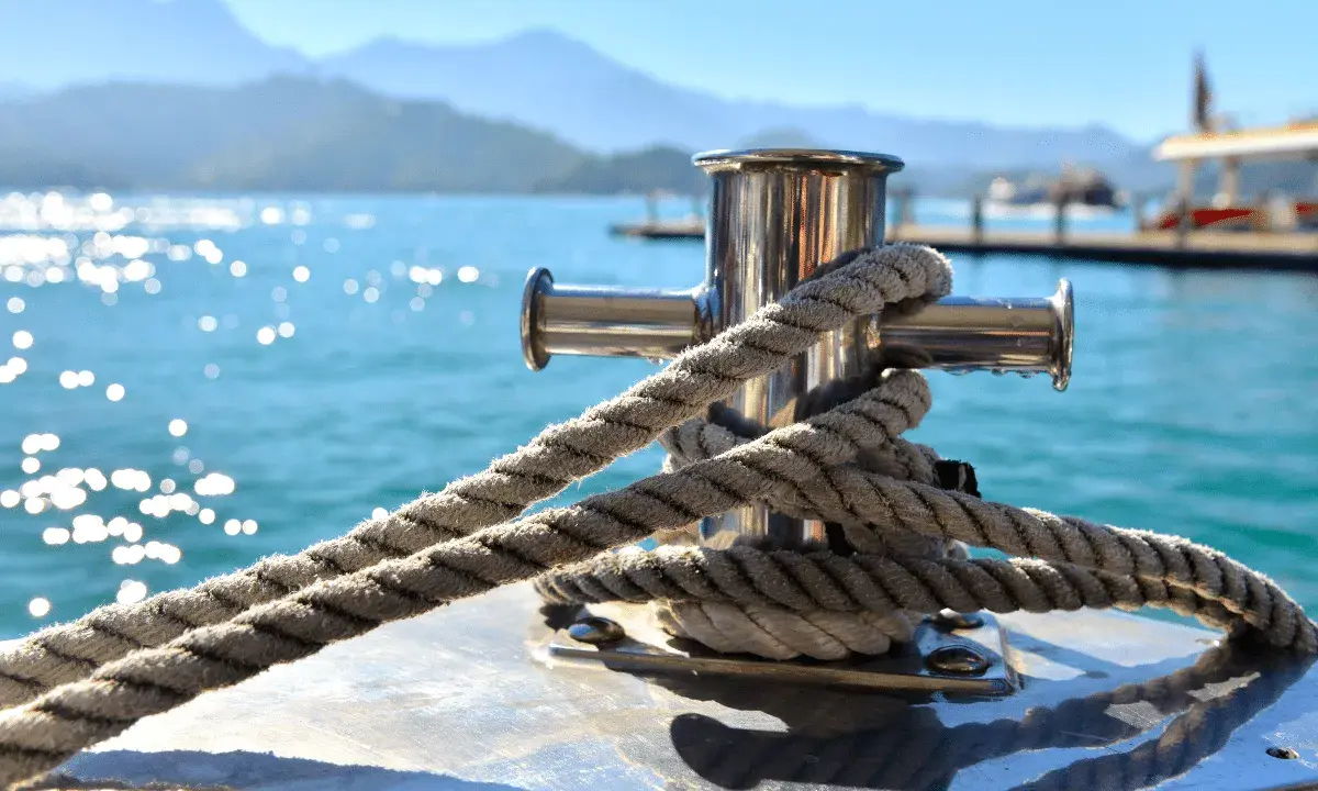 Gros plan sur une ligne de mouillage corps mort, une corde épaisse enroulée autour d'un taquet en acier inoxydable sur un bateau. L'eau bleue scintillante et les montagnes floues en arrière-plan créent une scène paisible.