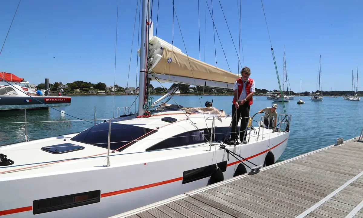 Un homme en gilet rouge ajuste une aussière sur un bateau blanc, prêt à appareiller. D'autres voiliers sont au mouillage.