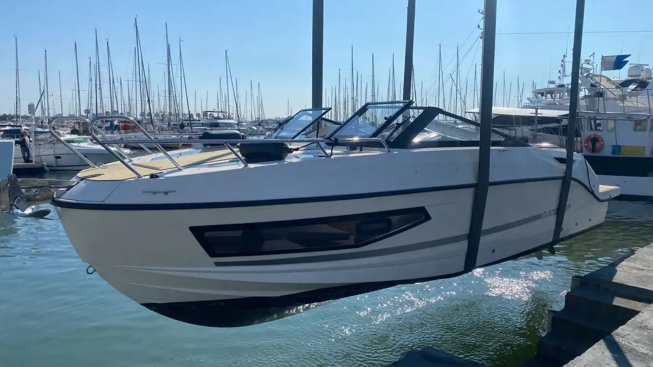 Un bateau blanc est soulevé par une grue, sa coque reflétant le soleil sur la surface mouillée du port.