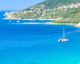 Un catamaran blanc avec des accents rouges navigue sur des eaux bleues cristallines, près d'une côte verdoyante et d'un village pittoresque.