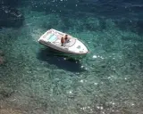Deux personnes dans un bateau blanc, au moteur en panne, dérivent sur une eau turquoise et claire, révélant les fonds rocheux.