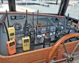 Plusieurs talkies-walkies sur un tableau de bord de bateau. Ils sont de différentes couleurs (orange, jaune, noir) et affichent des canaux. Une discussion sur la différence entre UHF et VHF pourrait être animée avec ces appareils.