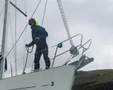 Un marinier, les amarres avis, manœuvre sur le pont d'un voilier blanc, face à une côte rocheuse verdoyante.