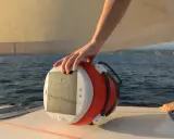 Main d'un marin tenant une bouée orange et blanche, un câble y est accroché. Le bateau navigue sur une mer bleue sous un ciel clair.