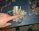 Main d'homme pointant un carburateur Zenith, possible cause d'un moteur 4 temps qui s'étouffe à l'accélération. Pièces de rechange visibles.