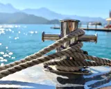 Gros plan sur une ligne de mouillage corps mort, une corde épaisse enroulée autour d'un taquet en acier inoxydable sur un bateau. L'eau bleue scintillante et les montagnes floues en arrière-plan créent une scène paisible.