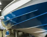 Un peintre en combinaison blanche applique une peinture bleue brillante sur la coque d'un bateau dans un atelier.