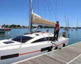 Un homme en gilet rouge ajuste une aussière sur un bateau blanc, prêt à appareiller. D'autres voiliers sont au mouillage.