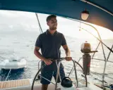 Un homme souriant est au gouvernail, prêt à barrer un bateau. Le soleil brille sur l'eau calme.