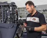 Un mécanicien effectue l'entretien d'un moteur hors-bord 4 temps Yamaha, tenant une bougie et un outil.