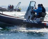 Deux bateaux Zodiac naviguent sur l'eau. L'un d'eux, au premier plan, porte le logo "ZODIAC" et semble communiquer sur le canal 9 VHF.