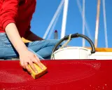 Maintenant, on apprend comment laver une coque de bateau. Une main savonneuse frotte la surface rouge avec une éponge jaune.