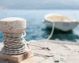 Gros plan sur un bollard en pierre avec une corde enroulée, prêt pour l'attache bateau pour quai. Un petit bateau est amarré en arrière-plan.