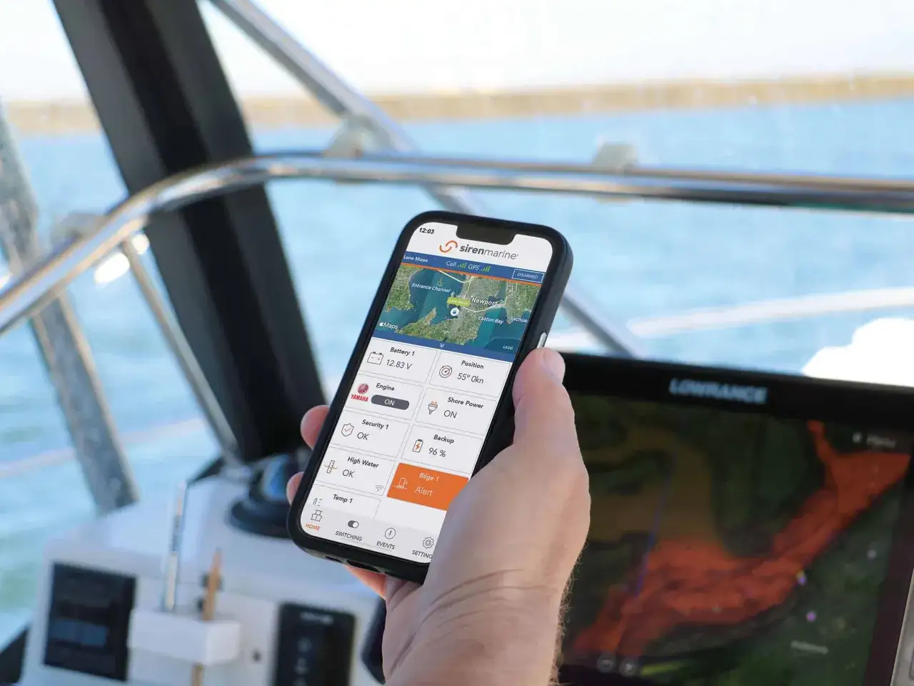 Main d'un plaisancier tenant un smartphone affichant une application de suivi de bateau. L'application affiche des informations sur la batterie, la position et l'état de la cale, permettant de surveiller la vitesse du bateau.
