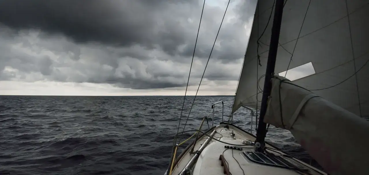Voile blanche sur mer agitée sous un ciel orageux. Le bateau navigue, prêt à affronter les éléments, une leçon sur comment prendre les vagues en bateau.