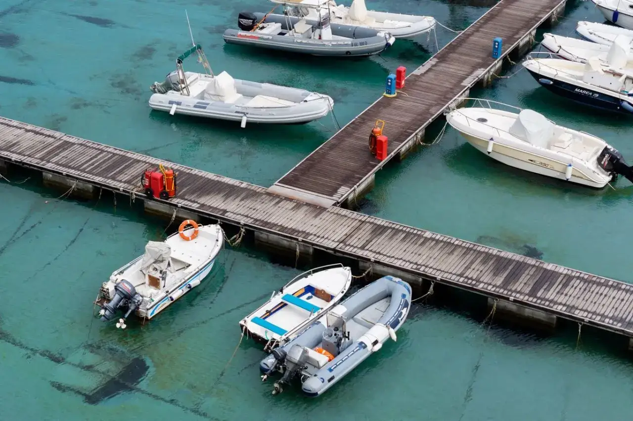 Plusieurs bateaux sont amarrés à un ponton en bois dans une eau turquoise. Des bidons rouges et bleus sont posés sur le ponton, prêts pour le prochain départ.