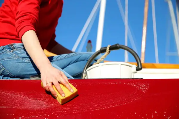 Maintenant, on apprend comment laver une coque de bateau. Une main savonneuse frotte la surface rouge avec une éponge jaune.